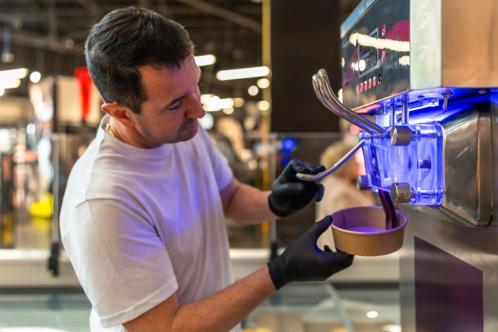 A gloved operator drawing soft serve into a paper cup from a commercial soft serve machine